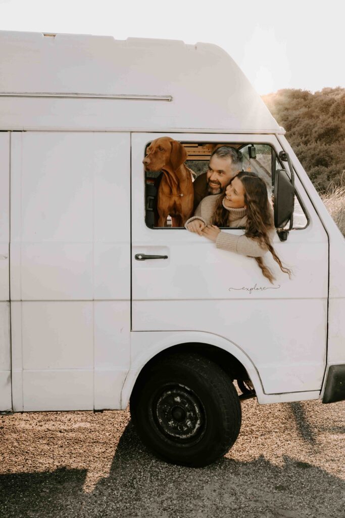 Ein brauner Hund lehnt aus einem weißen Camper-Van in Holland bei einem Hundefotoshooting, während ein Paar dahinter liebevoll lacht. Graslandschaft im Hintergrund.