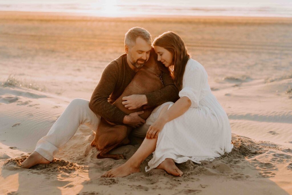 Familie mit braunem Hund am Strand bei Sonnenuntergang. Der Hund sitzt zwischen einer Frau im weißen Kleid und einem Mann in heller Kleidung.