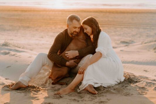 Familie mit braunem Hund am Strand bei Sonnenuntergang. Der Hund sitzt zwischen einer Frau im weißen Kleid und einem Mann in heller Kleidung.