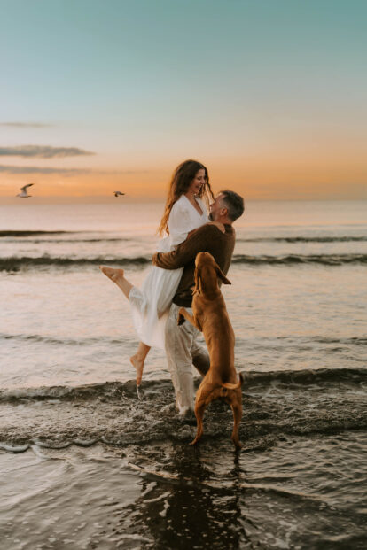 Ein Mann hebt eine Frau aus dem Wasser, während ein brauner Hund freudig zuschaut. Im Hintergrund: Strand und Möwen bei Sonnenuntergang