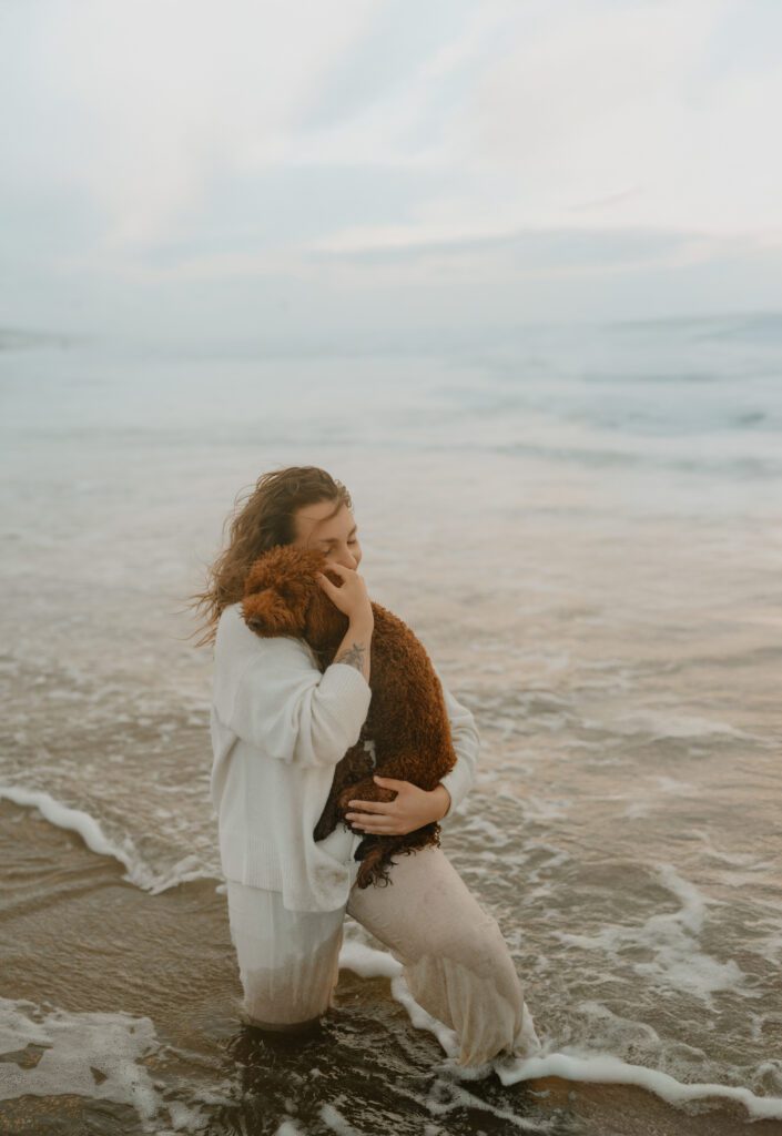 Frau mit langem, lockigem Haar lacht und hält ihren braunen Pudel am Wasser beim Hundeshooting in Holland.